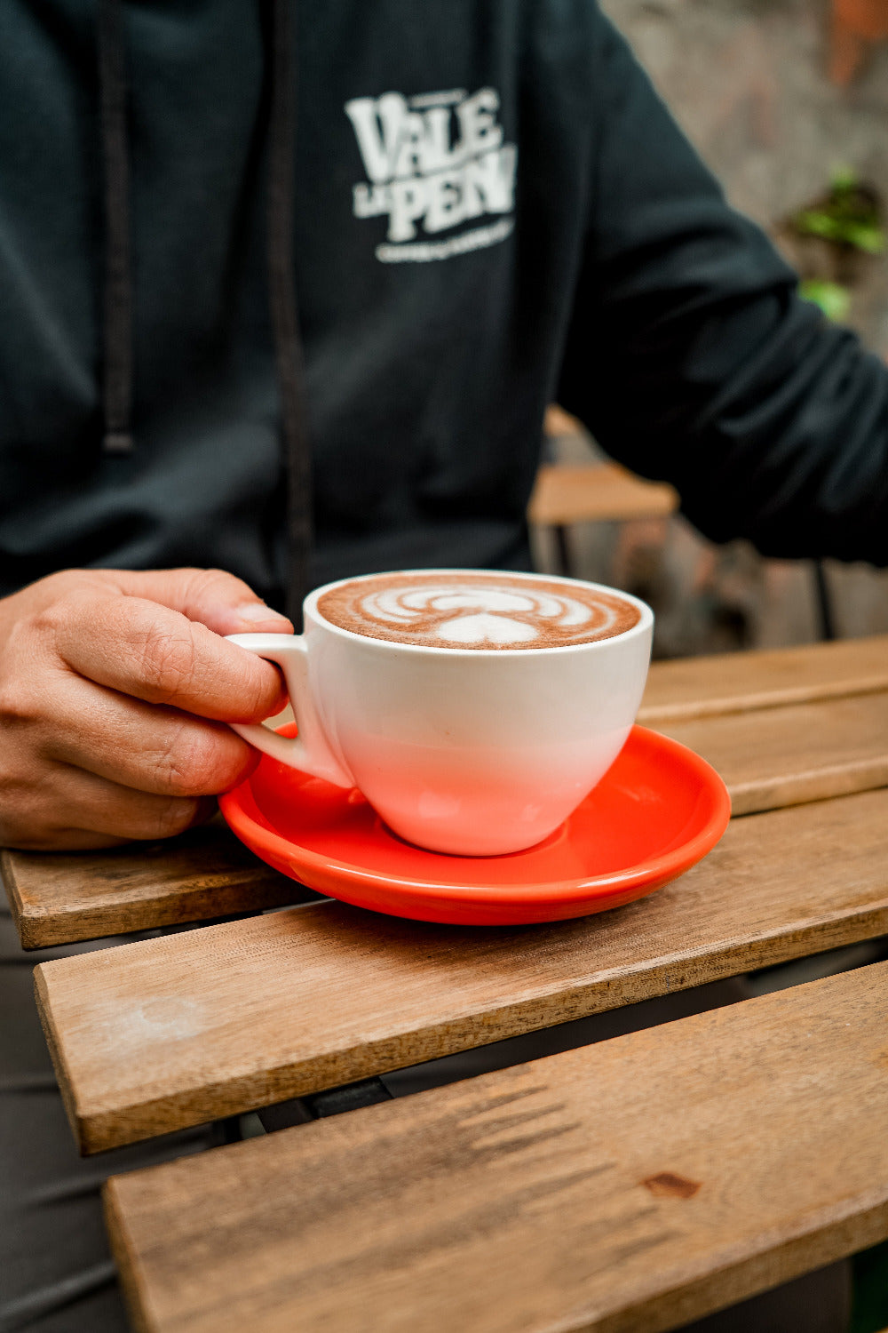 Person holding a white cup with a red saucer featuring a cappuccino on a wooden table.